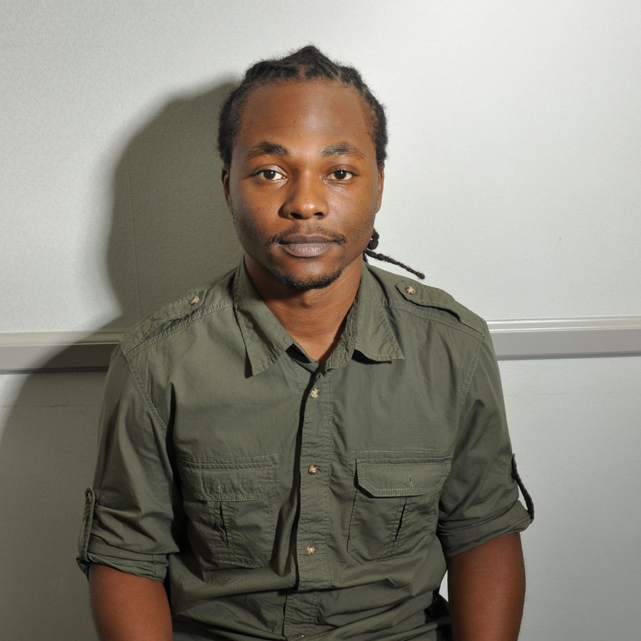 A young man sits behind bright lights casting shadows on the side of his head. The wall behind him has a thick, gray stripe.